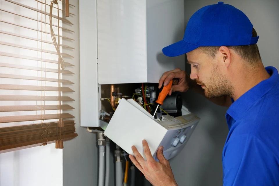 Technicien en uniforme bleu travaillant sur une chaudière murale domestique, consultant un manuel technique tout en effectuant une réparation avec un tournevis